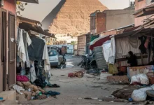 زقاق فقير في القاهرة يضم منازل متهالكة وملابس معلقة وفوضى، وخلفه تظهر أهرامات الجيزة العظيمة، يبرز التباين بين الفقر والعراقة التاريخية في مصر.