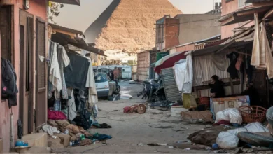 زقاق فقير في القاهرة يضم منازل متهالكة وملابس معلقة وفوضى، وخلفه تظهر أهرامات الجيزة العظيمة، يبرز التباين بين الفقر والعراقة التاريخية في مصر.
