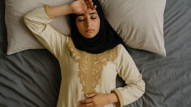 امرأة شابة من أصول شرق أوسطية ترتدي لباسًا تقليديًا مطرزًا وحجابًا أسود، مستلقية على سرير أبيض بنظرة متعبة، ملتقطة من زاوية علوية، تعبر عن الإرهاق العقلي في العصر الحديث